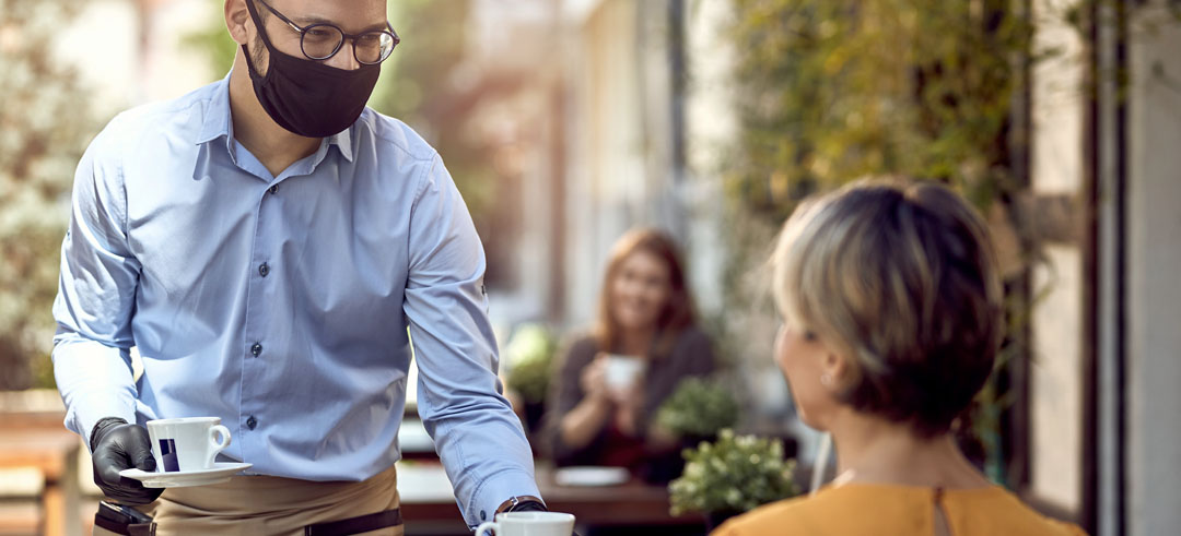 Camarero con mascarilla y guantes llevando dos cafés a clientas